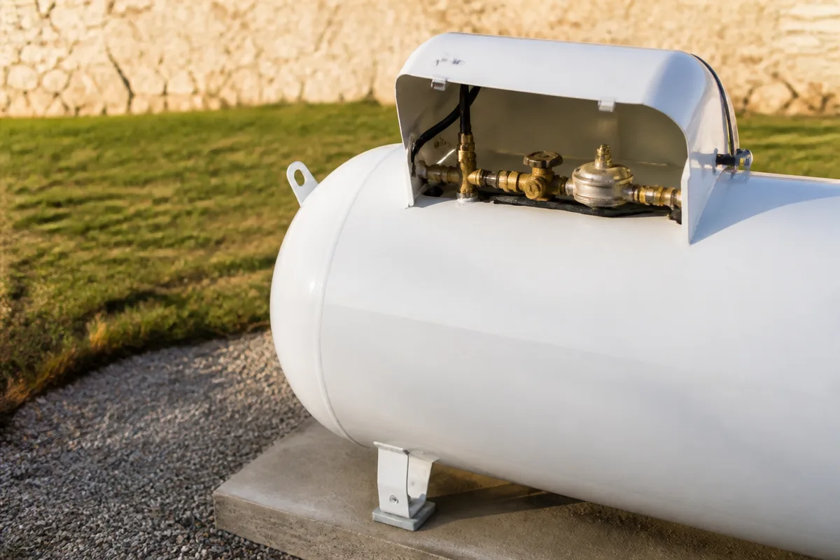 Citerne de propane aérienne posée sur un socle de béton derrière une maison de calcaire clair du Larzac